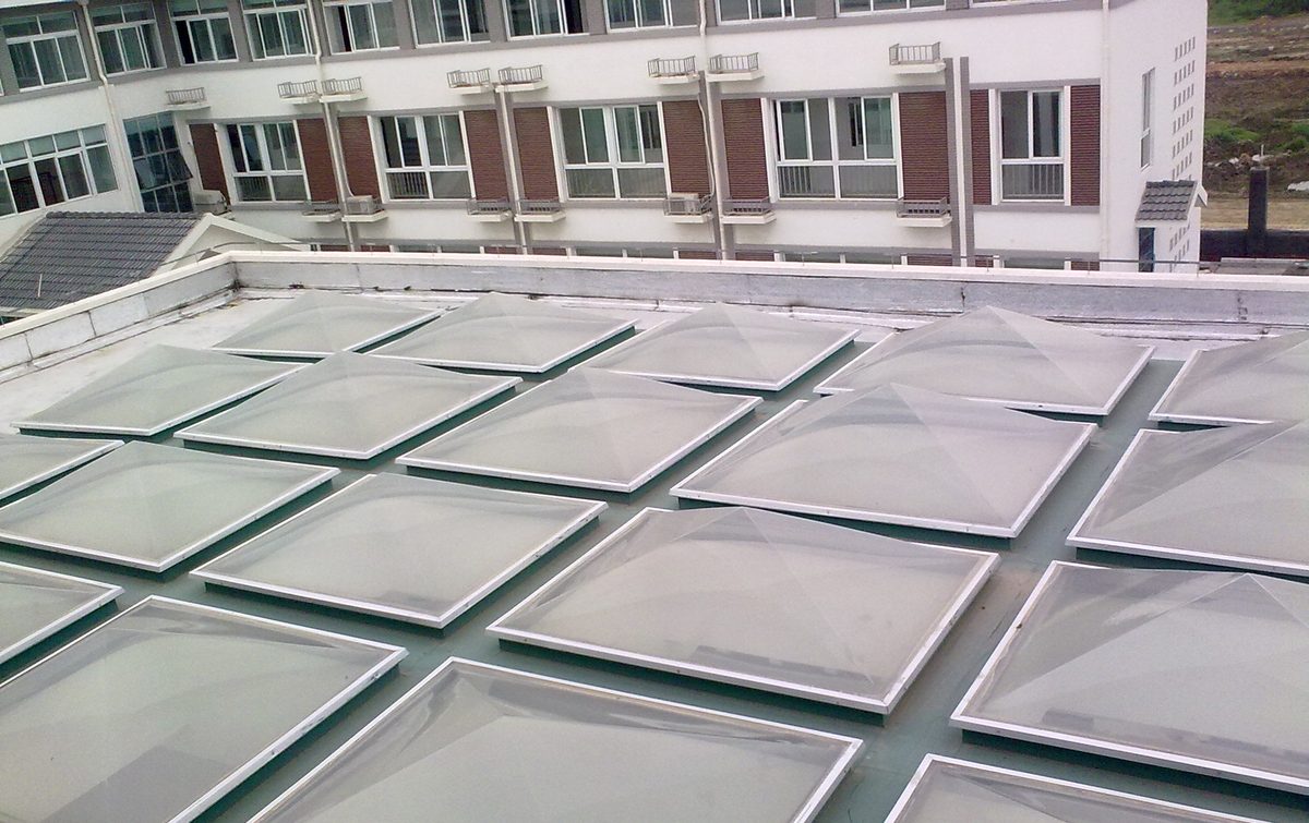 Grid of translucent polycarbonate pyramid skylight domes on a flat rooftop beside a multi-storey building